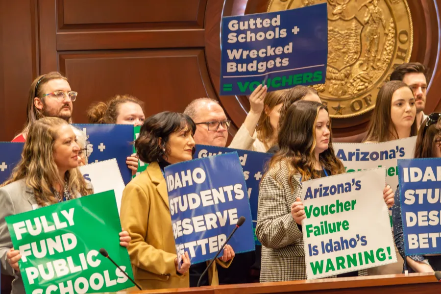 Protesters in a government chamber hold signs opposing vouchers and school cuts, including Fully Fund Public Schools.